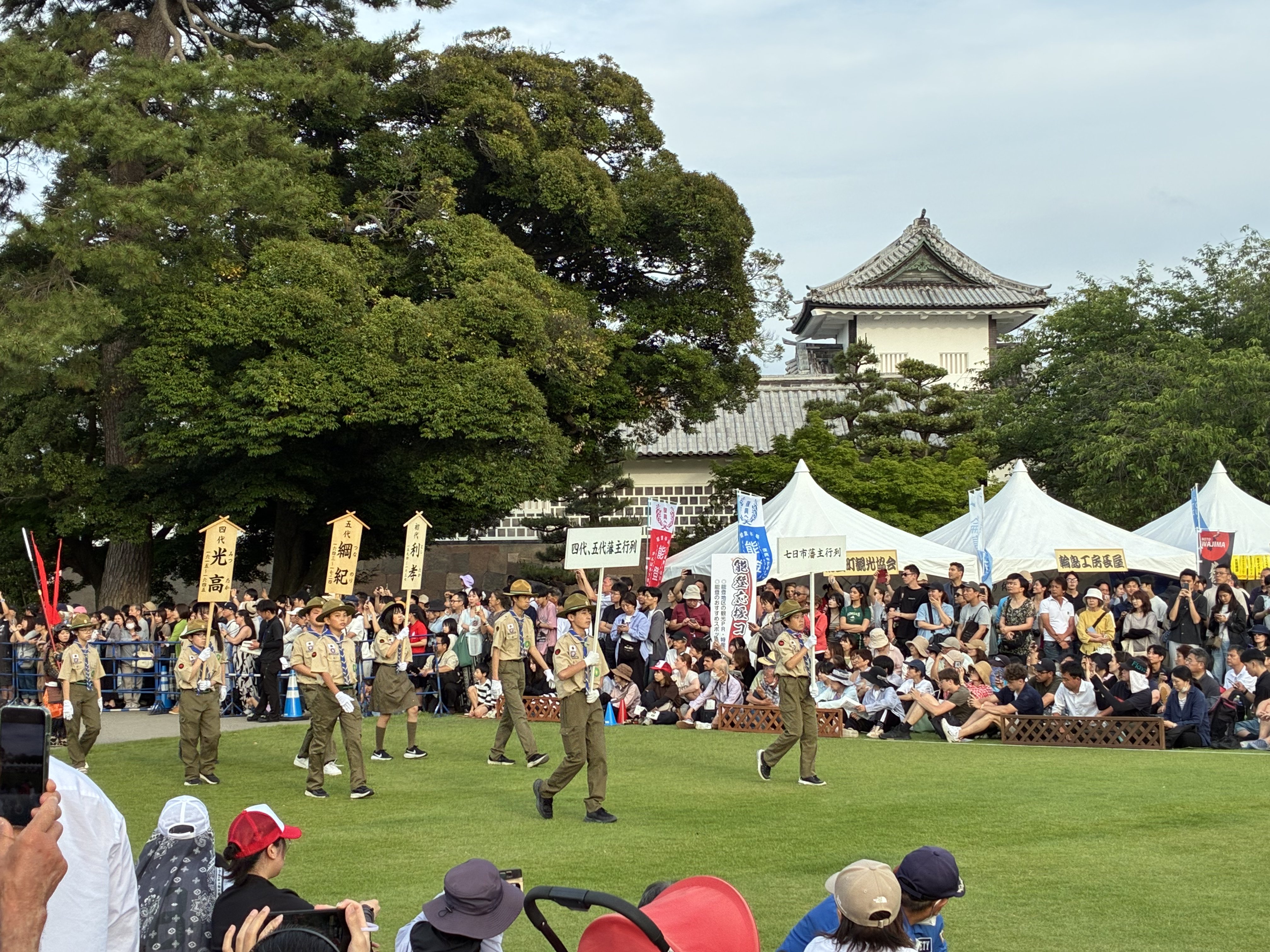 旗を持った茶色い衣装の行列と金沢城の天守